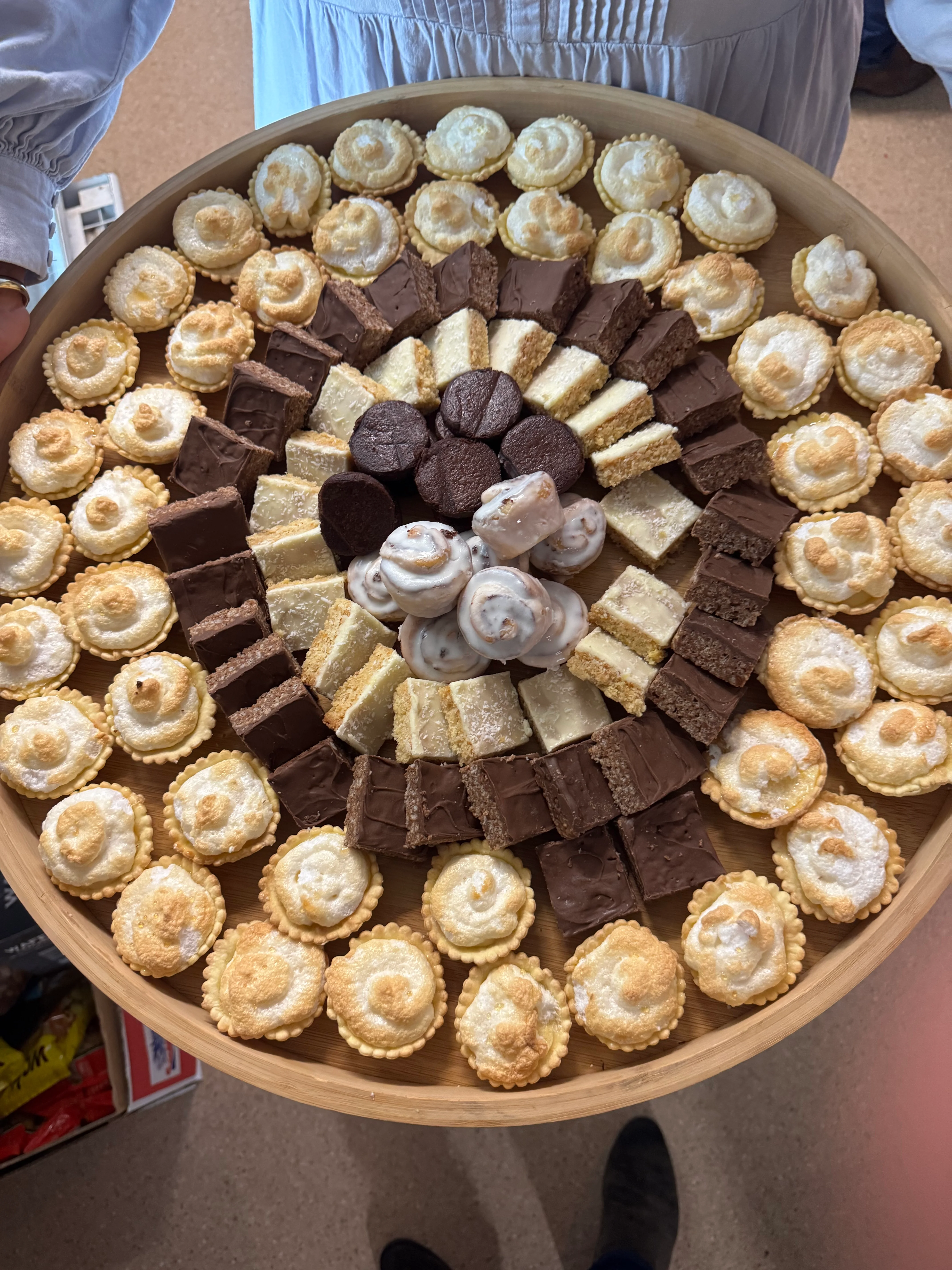 A wide round wooden platter of mini tarts, brownies, cream puffs and chocolate fingers.