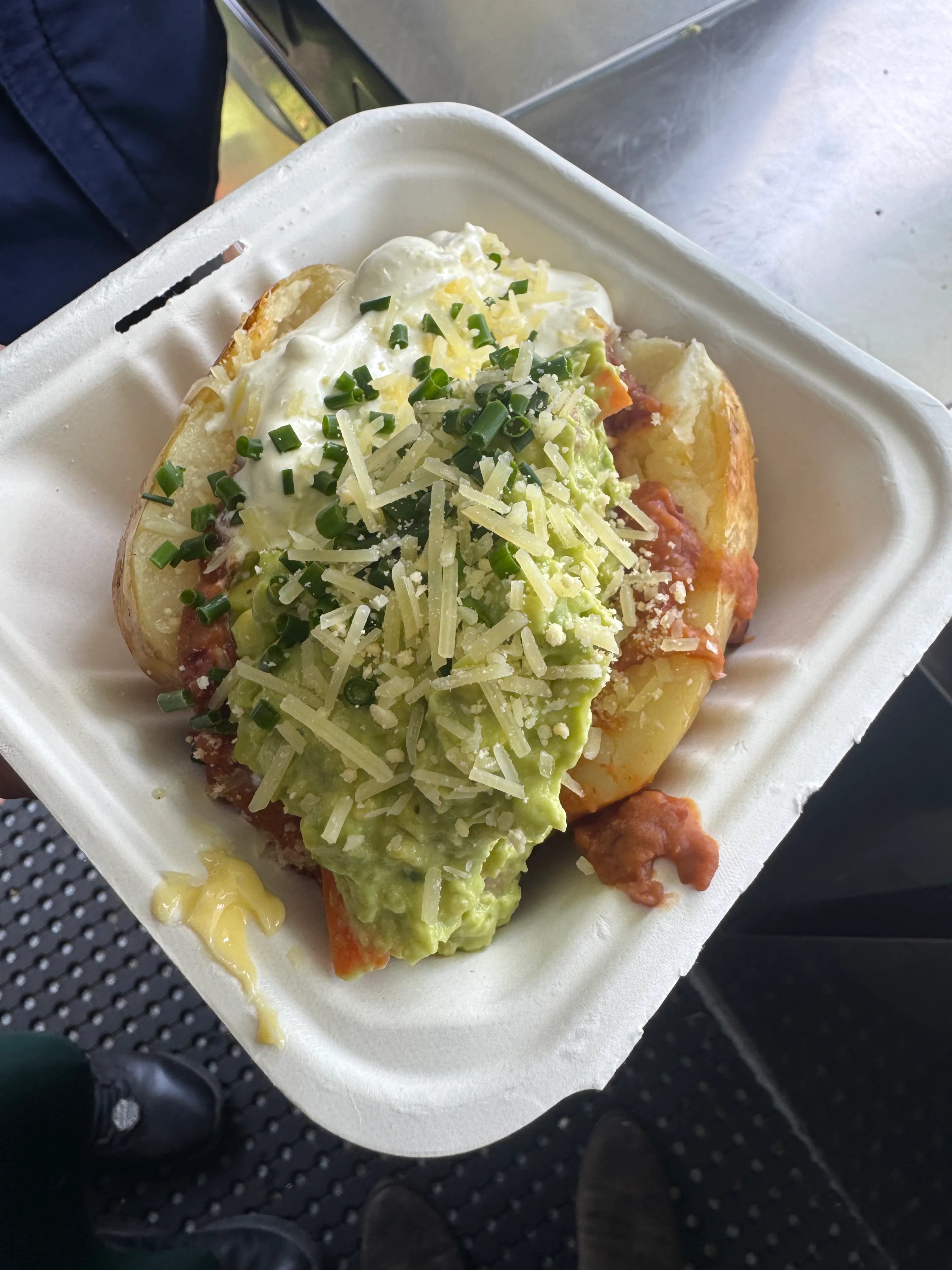 A loaded baked potato in a takeaway container — guacamole, sour cream, chives and shaved cheese.